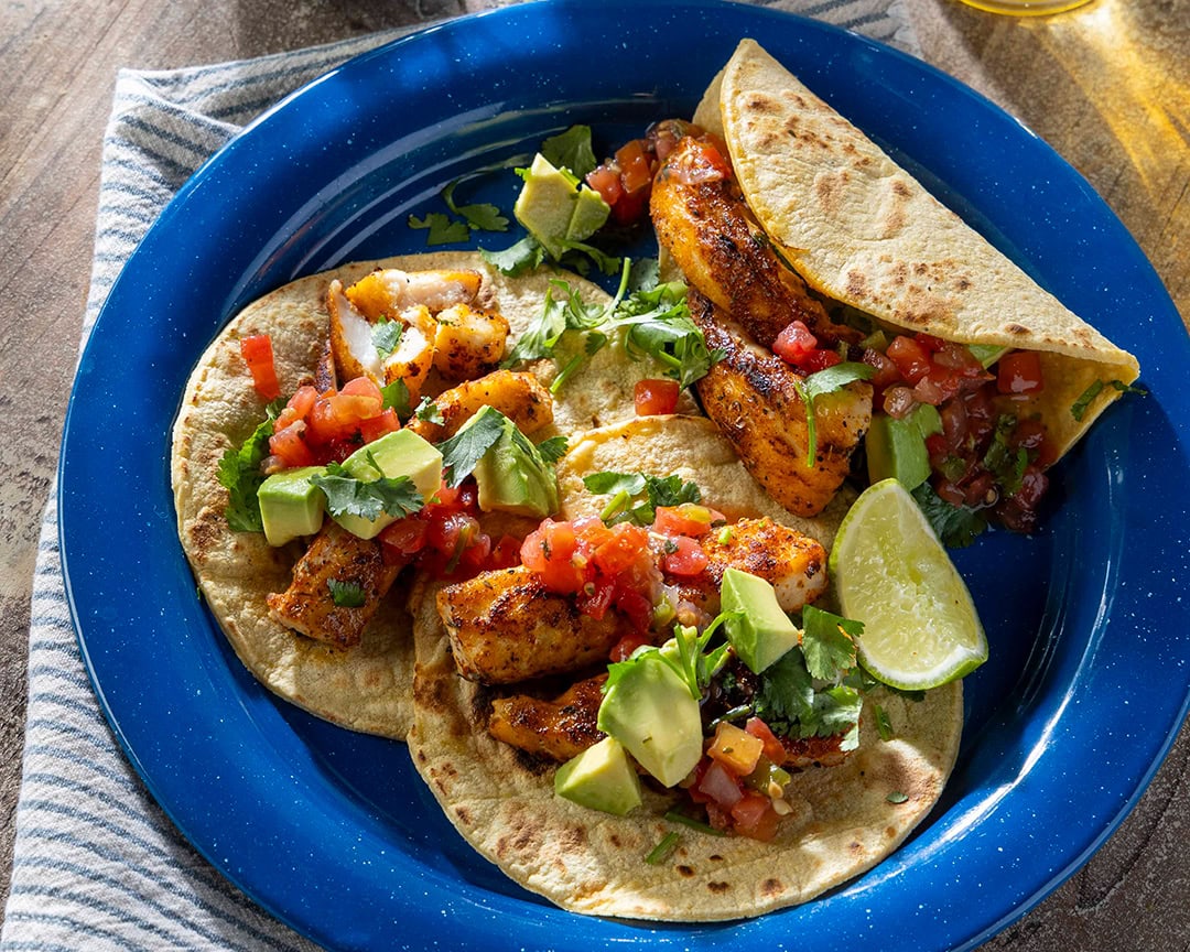 Three blackened fish tacos with lime and cilantro and diced tomato in a deep blue plate.