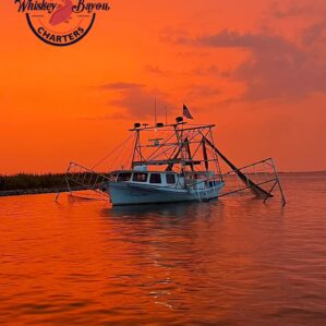 Image of a white fishing boat on the water. Both the sky and water are painted a orange red by the sunset.