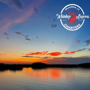 Calm marsh water in the morning light. The color is a black to blue to orange gradient. There are scattered clouds in the sky colored pink and orange. There is silhouetted land in the distance.
