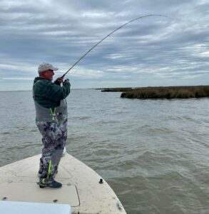 Man in camouflage overalls on overcast day fishing from the bow of a boat. Marsh is in the background.