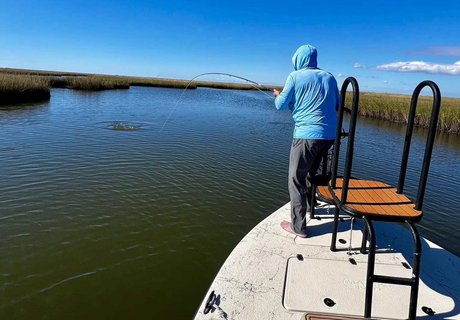 Man in blue hoodie fishing from the bow of a boat. Its a clear sunny day and the blue water and sky are broken up only by the tan reeds.