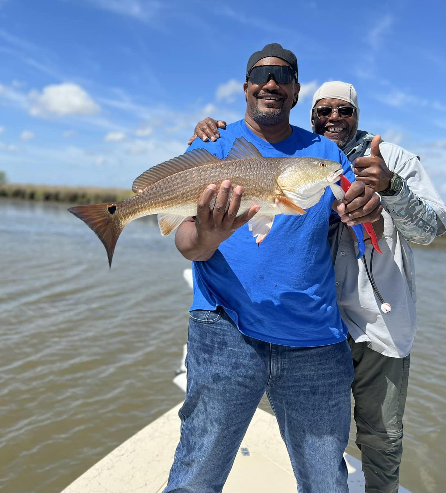 Once man proudly holding up his catch for the camera while his buddy stands behind him with one hand on his shoulder and the other giving a thumbs up