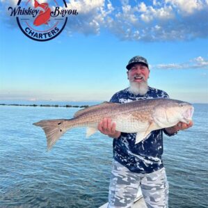 A smiling bearded man smiling from the prow of a boat as he holds up a large Redfish.