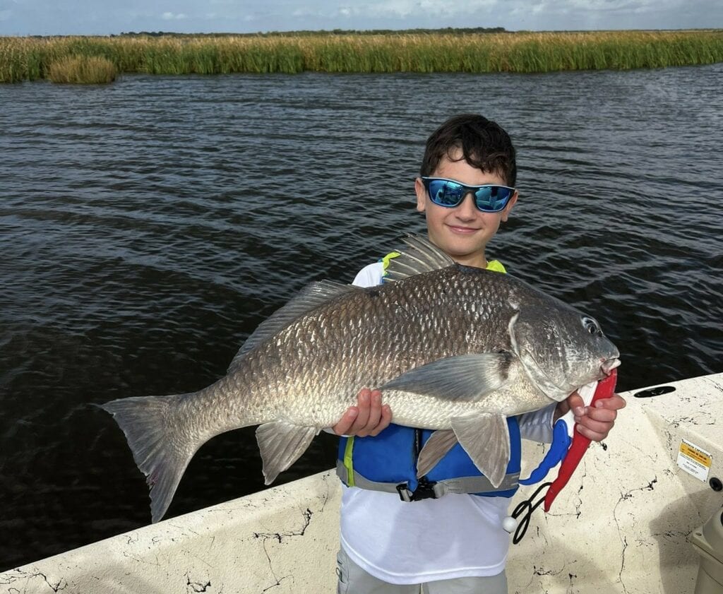 A young boy, sporting sunglasses and a life vest, holding up his big catch for the camera. Behind him is the high grass of the Louisiana marsh.