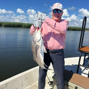 Man in a pink shirt standing in a boat holding up the fish he just caught.